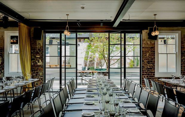 A refined restaurant dining room with long, fully set tables, rows of contemporary upholstered chairs, exposed dark brick walls, black-framed glazed doors and windows, and a covered courtyard visible beyond. Pendant lights and a white slatted ceiling create a polished industrial-contemporary atmosphere.