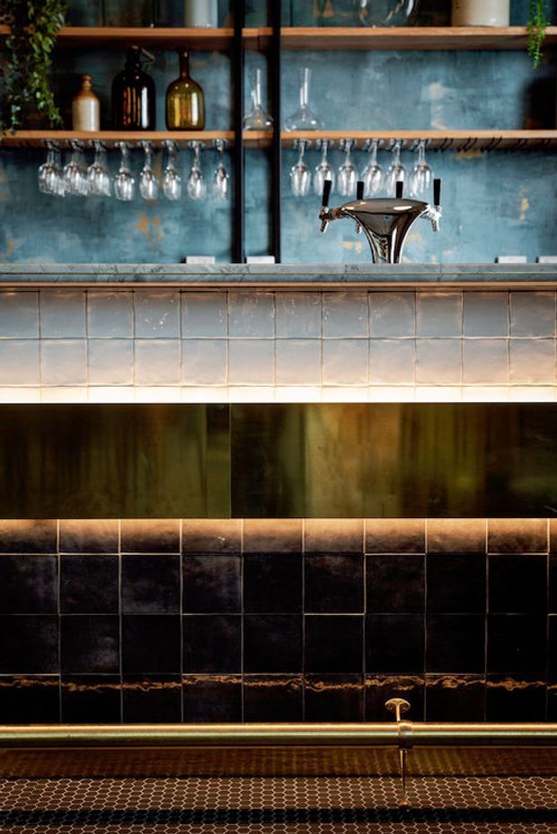 Close-up of a sophisticated bar counter with open timber shelves displaying bottles and hanging wine glasses, a beer tap, blue-grey textured backsplash, pale square tiled counter front, dark glossy tiled lower wall, and warm metallic trim and cabinetry. The composition emphasizes rich materials, layered lighting, and refined hospitality detailing.