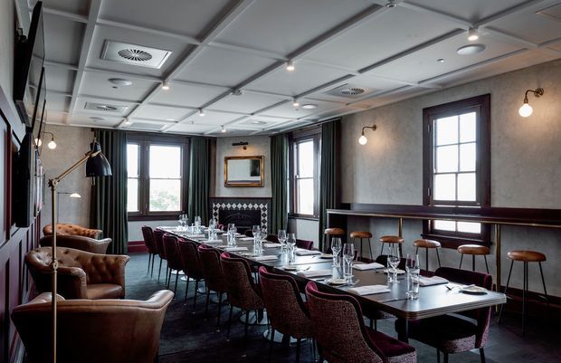 A refined dining and event room with a long formally set table, upholstered burgundy dining chairs, leather lounge chairs, dark timber bar-height ledge, tall multi-pane windows, and a coffered ceiling with recessed lighting. The palette is dark, warm, and sophisticated, with a small fireplace or decorative feature at the far wall.