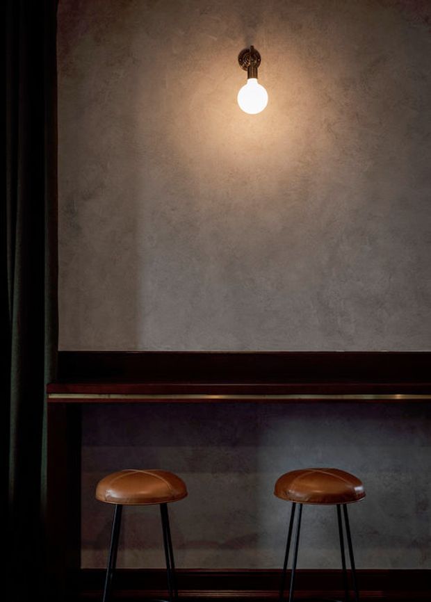 Moody close-up of a dark, refined interior featuring a textured plaster wall, a single exposed globe pendant light, dark timber framing and trim, a narrow counter or ledge with a brass-toned edge, and two brown leather bar stools with slender black metal legs.