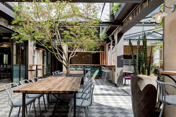 A stylish semi-enclosed courtyard dining area with a large reclaimed timber communal table, metal chairs, checkerboard floor tiles, mature indoor tree, built-in greenery, exposed dark structural beams, and extensive overhead glazing. The space has a refined hospitality or restaurant character with an open connection to adjacent interiors.