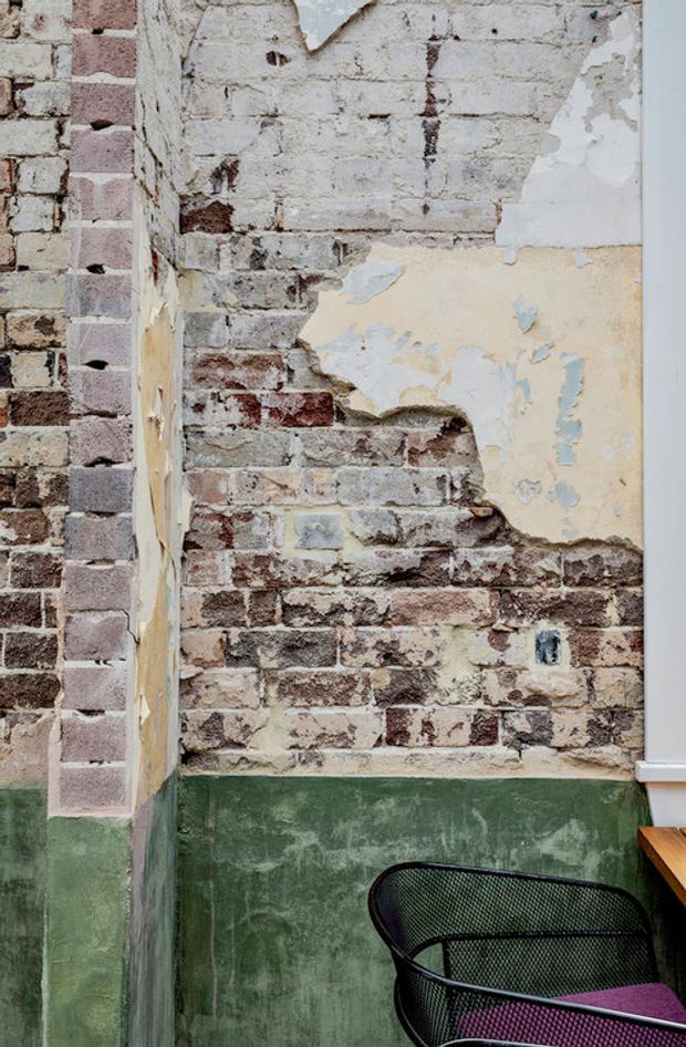 Close-up interior view of a distressed, historic brick wall with extensive peeling white and pale yellow plaster, exposed masonry, and a dark green painted lower section. A black metal mesh chair and the edge of a timber tabletop appear in the foreground, suggesting an informal commercial or hospitality setting. The image emphasizes layered, weathered textures and adaptive-reuse character.