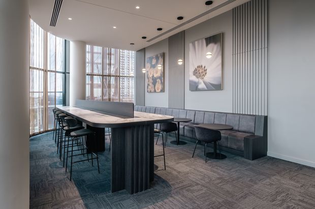 A contemporary communal dining and lounge area, likely in a commercial or hospitality setting. A long rectangular dining table with bar stools is paired with a built-in upholstered banquette and small round tables. Floor-to-ceiling glazing provides abundant daylight, while vertical wall panelling, pendant lights, recessed downlights, and restrained grey-blue finishes create a refined modern interior.