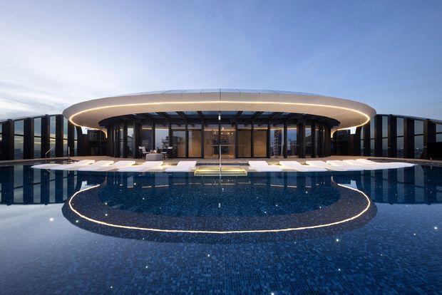 A dramatic dusk exterior view of a low-profile contemporary residence or hospitality-style building, fronted by a large reflective swimming pool. The building features extensive dark-framed floor-to-ceiling glazing, a broad curved projecting roof or canopy with integrated warm LED lighting, and symmetrical poolside lounge areas. The pool includes a dark blue tiled finish and a curved illuminated feature near the foreground.