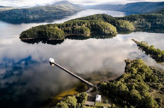 Aerial view of a tranquil lake surrounded by densely forested islands and mountainous terrain. A long narrow jetty extends from the shoreline to a small white structure on the water, with another small white building visible among the trees near the shore.