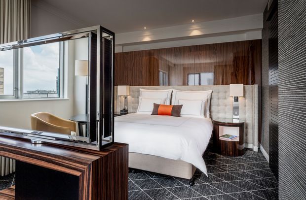 Luxurious contemporary bedroom featuring a large upholstered bed with layered white bedding, dark wood wall panelling and matching bedside tables, mirrored cabinetry or a dressing console in the foreground, and patterned dark carpet. Floor-to-ceiling windows and neutral curtains bring in daylight, while symmetrical bedside lamps and a bold orange accent cushion add refined contrast.