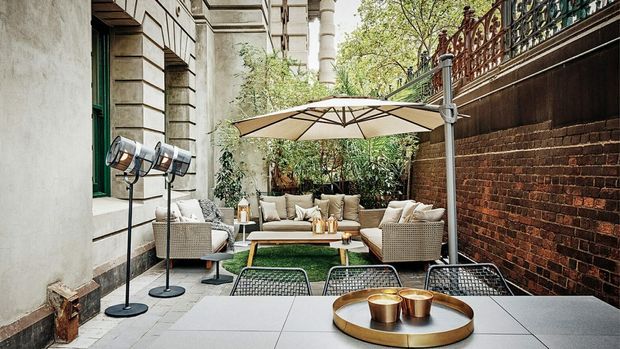 A refined urban courtyard terrace furnished as an outdoor lounge and dining area. The space features pale stone paving, a low outdoor sofa and armchairs, a central coffee table, a dining table in the foreground, a large cantilevered umbrella, studio-style floor lamps, a weathered brick wall, and ornate metal railing above. Mature planting softens the enclosed courtyard.