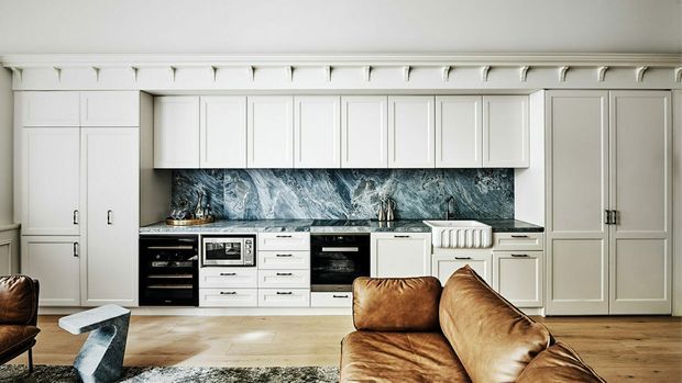 Bright, refined open-plan kitchen and living area with extensive white Shaker-style cabinetry, integrated appliances, a dramatic blue-grey marble-look splashback, pale timber flooring, and a brown leather sofa in the foreground. Decorative ceiling cornices and panelled cabinetry add a classic architectural character.