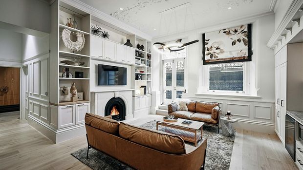 Bright, refined living room with built-in white cabinetry and open shelving, a central fireplace, wall-mounted television, leather sofas, a coffee table, and decorative wall panelling. Tall windows and glazed double doors bring in natural light, while ornate ceiling detailing and a framed botanical artwork add a traditional character.