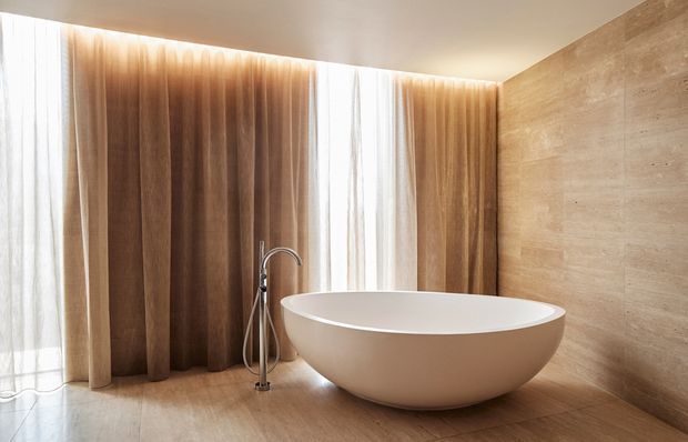 A serene contemporary bathroom featuring a freestanding oval bathtub, wall-mounted or floor-standing bath filler, full-height sheer curtains with warm timber-toned drapery, and beige stone or concrete-look wall and floor finishes. Soft concealed lighting creates a warm, luxurious atmosphere.