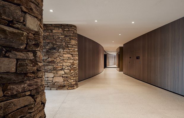 A wide, softly curved interior corridor with a pale polished concrete or terrazzo-like floor, warm dark vertical timber wall panelling, and dramatic rough stacked-stone cylindrical columns and walls. Recessed ceiling lights provide even illumination, while the sweeping curved geometry creates a refined, gallery-like circulation space.
