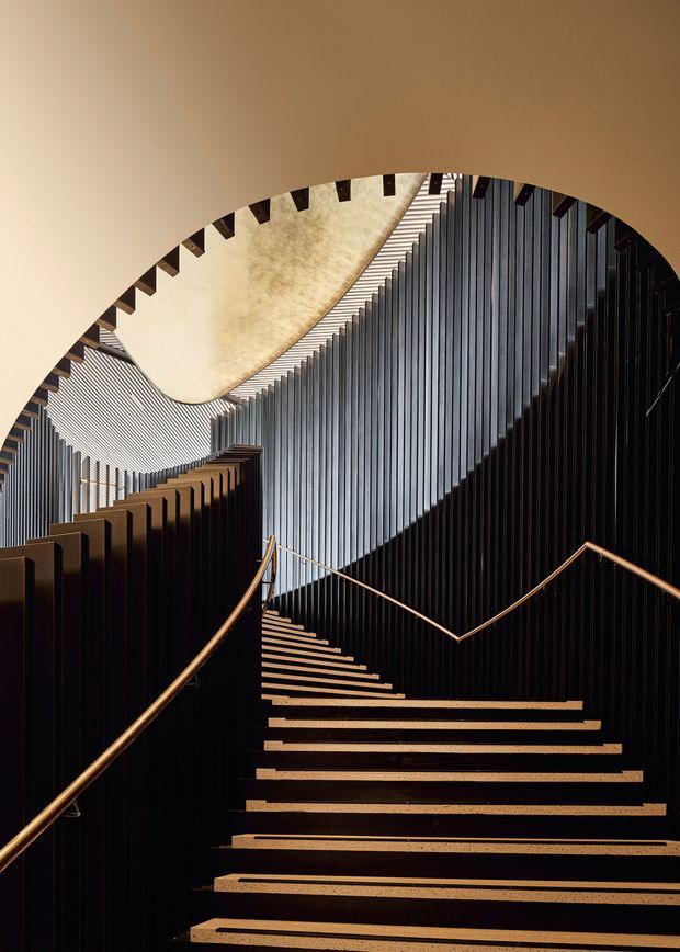 Dramatic interior architectural photograph of a sculptural stairway curving upward through a tall space. Broad dark stair treads are edged in warm wood or bronze tones, with slender bronze handrails and dense vertical black slatted wall elements. A large sweeping pale curved ceiling or canopy and strong directional lighting create a striking geometric composition.