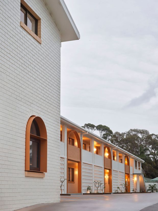 Exterior architectural photograph of a multi-storey building with pale painted brick walls, repeated arched windows and openings framed in warm timber, illuminated recessed balconies, and decorative breeze-block screens. A broad paved forecourt occupies the foreground, with mature trees beyond. The building has a long repetitive form and a shallow pitched roof with broad eaves.