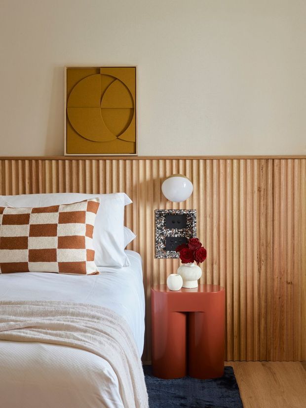 A warmly styled bedroom vignette featuring a partially visible bed with white bedding and a rust-and-cream checkerboard cushion, a vertical fluted timber feature wall, and a compact terracotta cylindrical bedside table. A sculptural wall light, decorative objects, red flowers, and geometric artwork add contemporary character. Timber flooring and a dark blue rug complete the composition.
