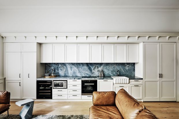 A refined white kitchen with extensive panelled cabinetry, integrated appliances, a dramatic blue-grey marble-look splashback, and a timber island or bench in the foreground. Decorative cornice detailing and small brackets add a traditional architectural character, while leather seating and pale timber flooring create a warm, contemporary living-kitchen setting.