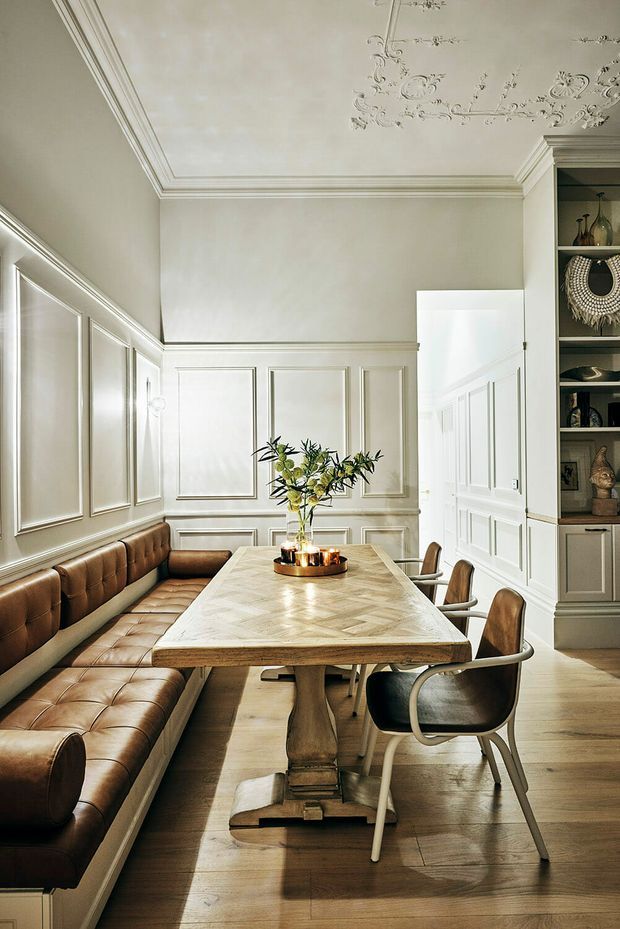 Elegant dining room with a long rectangular wood dining table, integrated tufted brown leather banquette seating, and coordinating dining chairs. The room features pale panelled walls, ornate decorative ceiling plasterwork, detailed cornices, built-in shelving, and warm timber flooring. Soft natural light creates a refined, welcoming atmosphere.