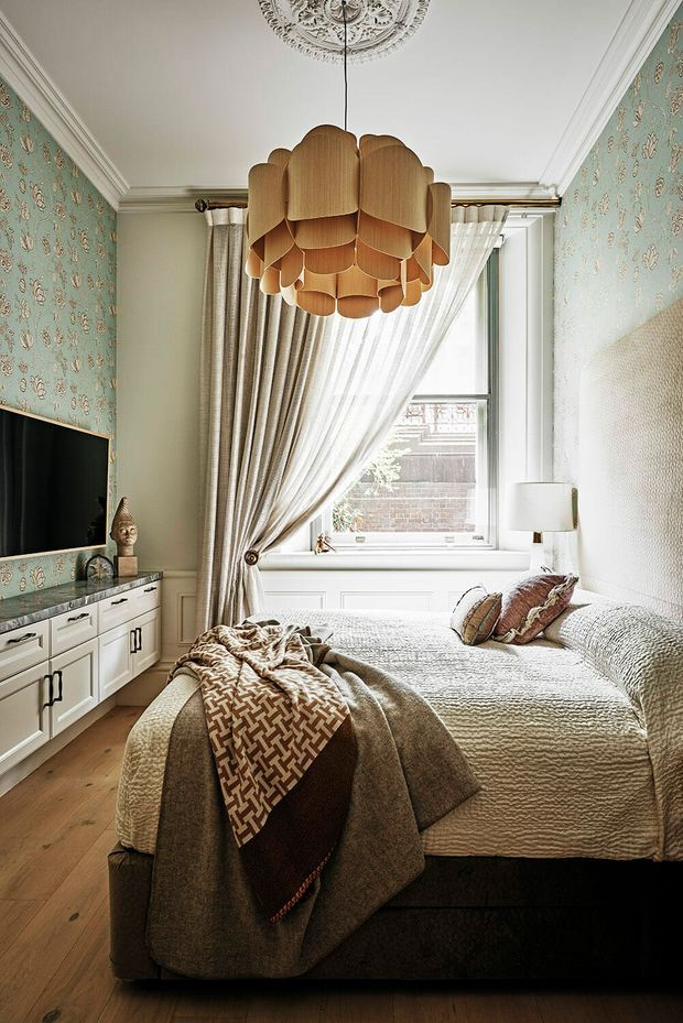 A richly styled bedroom with a large upholstered bed, layered neutral bedding and throws, patterned blue-green wallpaper, white built-in cabinetry, and a dramatic sculptural wood pendant light. Tall curtained glazing brings in natural light, while ornate ceiling detailing and refined traditional trim add character.