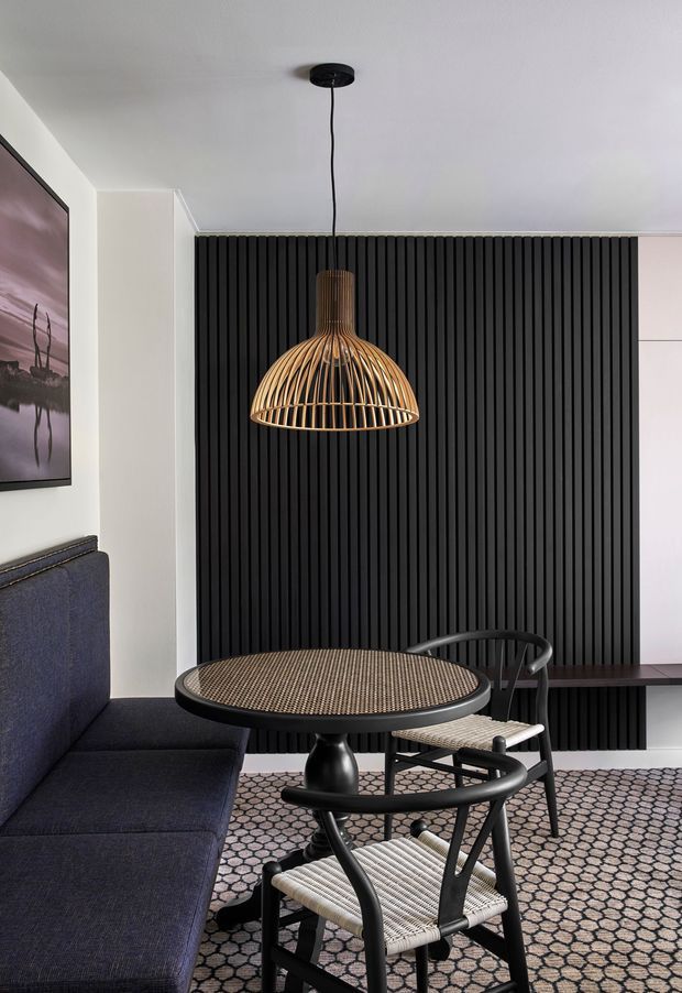 A refined dining and living area with a dark upholstered banquette, round dining table, woven black-framed chairs, patterned rug, and a sculptural woven pendant light. A dramatic black vertical slatted feature wall contrasts with pale walls and ceiling, while artwork and integrated cabinetry complete the contemporary interior.