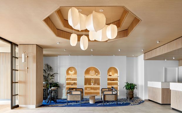 A refined contemporary lobby or reception area with warm timber detailing, vertical slatted wall panelling, arched illuminated display niches, lounge seating, plants, and a striking sculptural pendant installation set into a recessed timber ceiling feature. A white reception counter occupies the right side, while blue patterned carpeting adds contrast to the neutral and wood-toned palette.