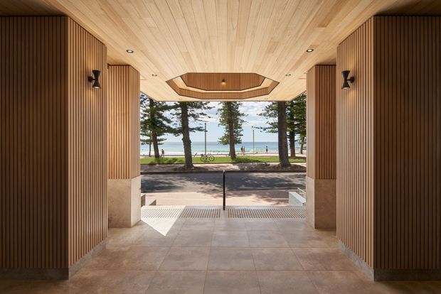 A refined covered entrance or passage framed by warm vertical timber wall slats and a matching timber-lined ceiling. Large open glazing or an open end reveals a landscaped waterfront setting with trees, a promenade, bicycles and the sea beyond. The floor is finished in large pale stone or concrete-look tiles, with discreet recessed lighting and dark wall sconces. A central geometric ceiling recess adds a sculptural architectural feature.