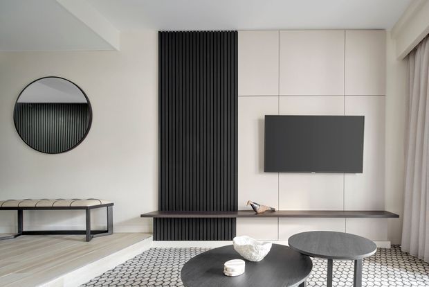 A refined contemporary living room featuring a wall-mounted television integrated into pale full-height cabinetry, a dramatic black vertical slatted feature panel with a projecting shelf, round black coffee tables, a geometric rug, a circular wall mirror, and expansive curtained glazing.