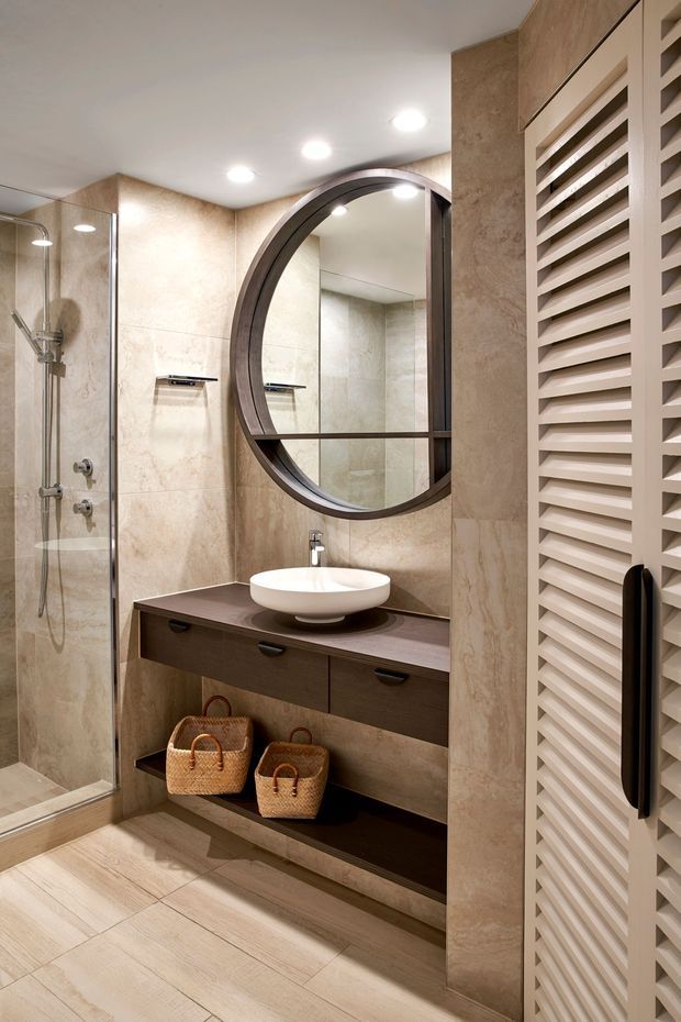 A compact, refined bathroom featuring warm beige stone-look wall and floor finishes, a frameless glass shower, floating dark timber vanity with a white vessel basin, and a large round dark-framed mirror. A tall white louvred cabinet door occupies the right side, while recessed ceiling lights provide even illumination.