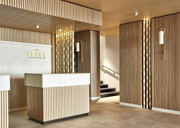 A refined hotel or club reception lobby featuring a fluted timber reception desk, matching vertical timber wall and ceiling slats, decorative metallic circular detailing, white textured counters, wall sconces, and a tiled floor. A sign identifies the space as The Sebel reception club. A stairway is visible beyond the lobby.