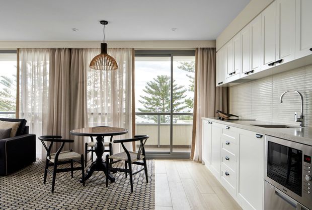 Bright open-plan kitchen and dining area with white shaker-style cabinetry, a pale tiled splashback, stone-look benchtop, stainless steel appliances, and a small round dining table with four black-framed chairs. Full-height glazing and sheer curtains open toward a balcony or exterior view, while patterned carpet and a dark sofa define the adjacent living area. A woven pendant light adds warmth.