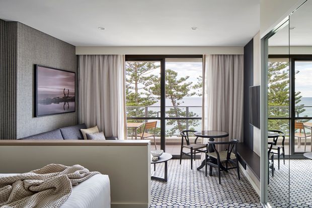 A refined open-plan bedroom and sitting area with a partial-height divider, overlooking a balcony and coastal landscape through large windows. The room features a grey upholstered sofa, textured grey feature wall, soft floor-to-ceiling curtains, a small black dining table with chairs, patterned carpet, and full-height mirrored cabinetry or glazing reflecting the seating area.