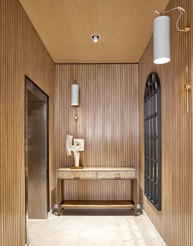A refined narrow entry hall featuring vertical timber wall panelling and a matching timber-lined ceiling, a slim console table with drawers, sculptural artwork, decorative wall lights, a dark arched internal window or glazed partition, and pale stone floor tiles. The space has warm, layered lighting and a sophisticated contemporary feel.