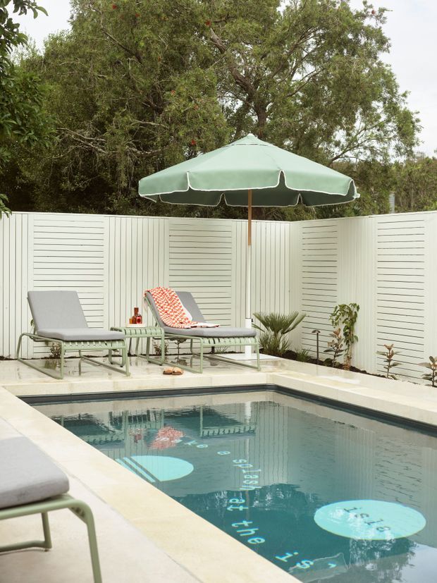 A private outdoor swimming pool surrounded by pale stone or concrete paving, white horizontal privacy fencing, and mature trees. Two pale green metal-framed sun loungers sit beneath a large mint-green market umbrella, with small potted and planted greenery around the pool. The pool water includes visible lane-style markings and playful circular graphics, creating a relaxed retro outdoor leisure setting.