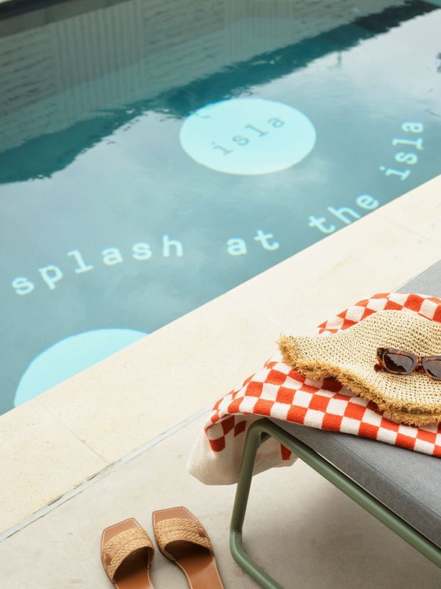 A styled poolside scene viewed from above, with turquoise swimming-pool water featuring large white lettering, pale concrete paving, and a green-framed lounge chair. The chair is draped with a red-and-white checkerboard textile and a woven straw hat with sunglasses, while tan sandals sit on the ground nearby. Bright, relaxed resort-like atmosphere.