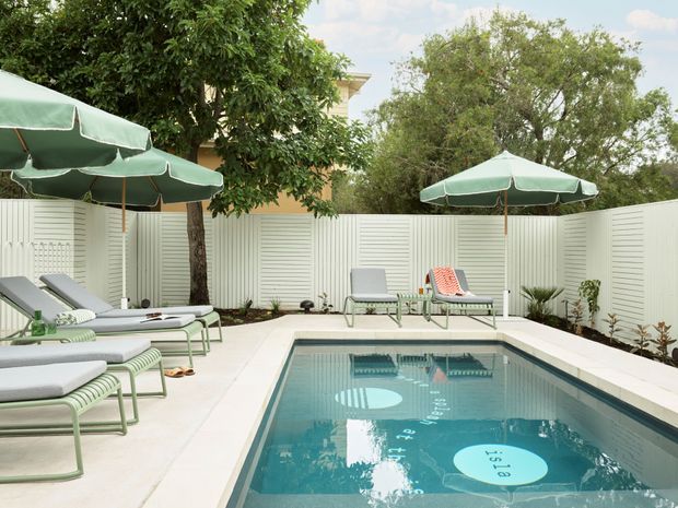 A private outdoor swimming pool courtyard with pale paving, a white slatted privacy fence, green market umbrellas, and coordinated striped pool loungers. Mature trees and clipped planting surround the pool, creating a relaxed mid-century-inspired resort atmosphere.