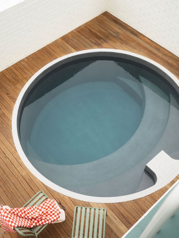 Top-down view of a compact oval plunge pool or spa with pale blue water, set into a warm timber deck. The pool has a light tiled rim and grey interior finish, with white painted brick walls enclosing the space. Two striped outdoor chairs and a red-and-white checkered towel appear at the edge of the deck.