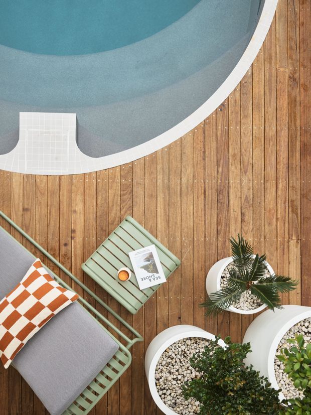 Top-down view of a landscaped outdoor pool deck with a curved swimming pool, warm timber decking, pale tiled pool edging and entry step, a green metal lounge chair with grey cushion, patterned orange-and-white pillow, side table, potted plants and a small book. The composition feels like a styled residential outdoor relaxation area.