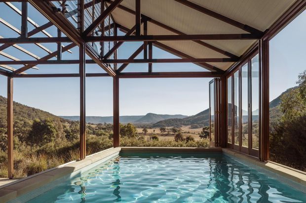 A covered indoor-outdoor swimming pool pavilion with extensive floor-to-ceiling glazing, exposed dark steel framing and roof trusses, and a pale ribbed ceiling. The pool occupies the foreground, while the open glazed sides frame a rural valley and rolling hills beyond.