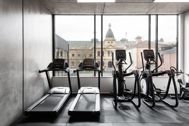 A modern commercial fitness room with treadmills, elliptical machines and exercise bikes arranged against a full-height glazed wall. The space has a raw industrial appearance with polished concrete ceiling and walls, dark tiled flooring, and large black-framed windows overlooking a historic urban building with a domed roof.