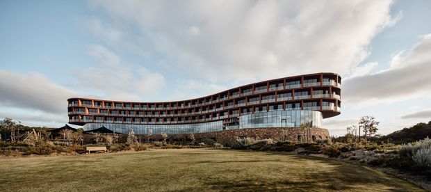 Wide exterior view of a large, gently curving multi-storey building set within landscaped grounds. The building has extensive repetitive glazing, dark horizontal facade bands, projecting balcony-like elements, a rounded end, and a substantial stone-clad lower level. The foreground is an open lawn with low planting, under a partly cloudy sky.