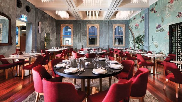 A richly decorated upscale restaurant dining room with multiple round and rectangular dining tables, red upholstered chairs, formal table settings, dark timber flooring, ornate textured walls, floral botanical wallpaper, framed mirrors, and layered geometric ceiling lighting. The space has a dramatic, luxurious and eclectic character with strong red, teal, grey and dark wood tones.
