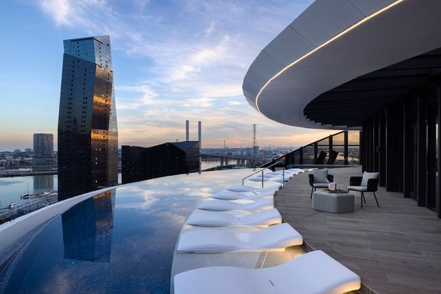 A high-end rooftop terrace overlooking an urban waterfront, with a curved infinity-edge pool, sculptural white sun loungers, and a covered outdoor seating area beneath a sweeping contemporary canopy. The scene includes a dark glazed high-rise and industrial waterfront structures in the background at dusk.