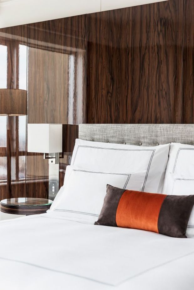 A refined bedroom featuring a neatly made bed with white linens and piped detailing, a grey upholstered headboard, and a bold orange-and-brown lumbar cushion. The bed is backed by a dramatic dark timber feature wall with glossy vertical grain, complemented by reflective timber wall panels, integrated bedside controls, a round dark timber side table, and modern table lamps. The overall composition is polished and luxurious with strong wood panelling and warm accent colour.