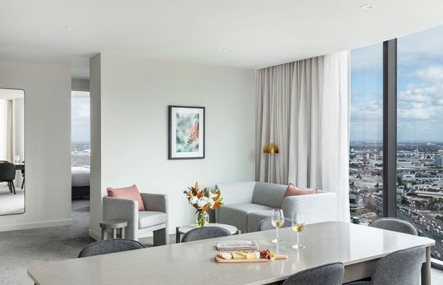 Bright, contemporary open-plan apartment interior combining a dining area and living room. A large pale stone island or dining bench with upholstered seating occupies the foreground, while a grey sofa, armchairs, coffee table, artwork, curtains, and floor-to-ceiling corner glazing frame expansive city views. The space has white walls, a light neutral palette, recessed ceiling lights, and a polished high-rise feel.