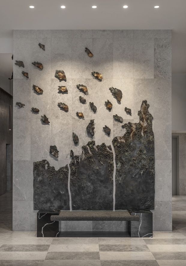 A dramatic contemporary interior feature wall clad in large pale grey stone panels, displaying an abstract dark metallic sculptural installation resembling an erupting or fragmented landscape. A slim built-in bench with dark veined stone supports sits at the base. Recessed ceiling spotlights illuminate the artwork, while matching large-format stone flooring extends across the surrounding lobby or entry space.