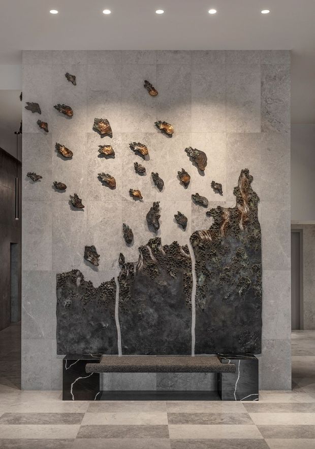 A refined interior feature wall in a lobby or entry space, clad in large pale grey stone-look panels and incorporating a dramatic dark sculptural artwork resembling an abstract landscape or cascading rock formation. A slim dark stone bench sits below the artwork, with matching dark stone pedestal ends featuring white veining. The surrounding floor is arranged in a large light and mid-grey checkerboard tile pattern, and recessed ceiling lights provide even illumination.