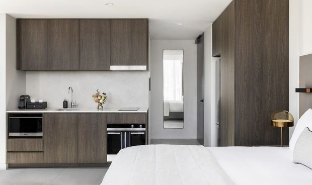 Compact open-plan studio-style interior with a bedroom in the foreground and a linear kitchenette along the left wall. The kitchen features dark wood-grain floor-to-ceiling and wall cabinetry, a pale stone or composite benchtop, integrated appliances, a stainless sink, and a light neutral backsplash. A narrow hallway with a full-length mirror leads toward a bright window, while the bed, white bedding, bedside surface, and brass-toned lamp occupy the foreground. The overall design is clean, modern, and space-efficient with a restrained neutral palette.