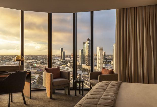 A refined high-rise bedroom or hotel-style suite with expansive curved floor-to-ceiling glazing overlooking a dense urban skyline at sunset. The room features a large upholstered bed in the foreground, two sculptural lounge chairs with a small round side table, a dark desk and chair near the window, full-height taupe curtains, warm neutral finishes, and soft ambient lighting. The panoramic glazing and elevated city view are the dominant architectural features.