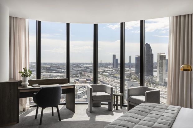 A refined high-rise bedroom with floor-to-ceiling curved glazing overlooking a dense city skyline. The room includes a dark built-in desk and upholstered chair, two light-grey lounge chairs with a small side table, a partially visible quilted bed, full-height curtains, and a brass-toned floor lamp. The palette is neutral and contemporary, with abundant daylight and expansive urban views.