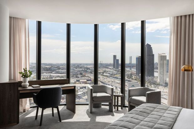 Bright contemporary high-rise bedroom with floor-to-ceiling curved glazing overlooking a dense urban skyline. The room includes a dark built-in desk and chair, two upholstered lounge chairs, a small side table, full-height curtains, and a partially visible quilted bed. The palette is neutral with dark timber accents and abundant daylight.