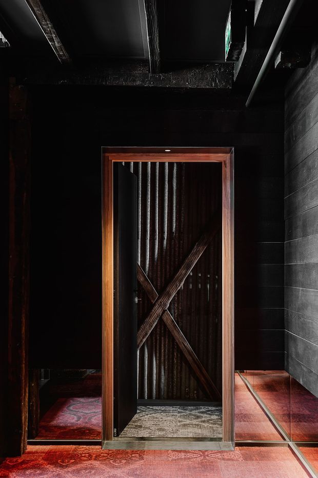 A dark, dramatic interior passage or entry area centred on a narrow timber-framed door. The door has a corrugated metal panel and prominent diagonal timber bracing forming an X, with black-painted walls and ceiling beams, a dark masonry or block wall to the right, and richly patterned red flooring extending through the space. The composition emphasizes rustic-industrial materials, strong contrast, and moody lighting.