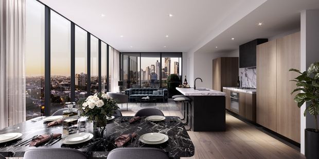 A high-end open-plan apartment interior combining a dining area, kitchen, and living room. The foreground features a dark marble dining table with upholstered dining chairs, tableware, and a floral arrangement. A compact kitchen with a dark island, waterfall-style benchtop, integrated appliances, and tall timber-toned cabinetry occupies the right side. Floor-to-ceiling black-framed glazing provides expansive city views, while recessed ceiling lighting and pale timber flooring create a refined contemporary atmosphere.